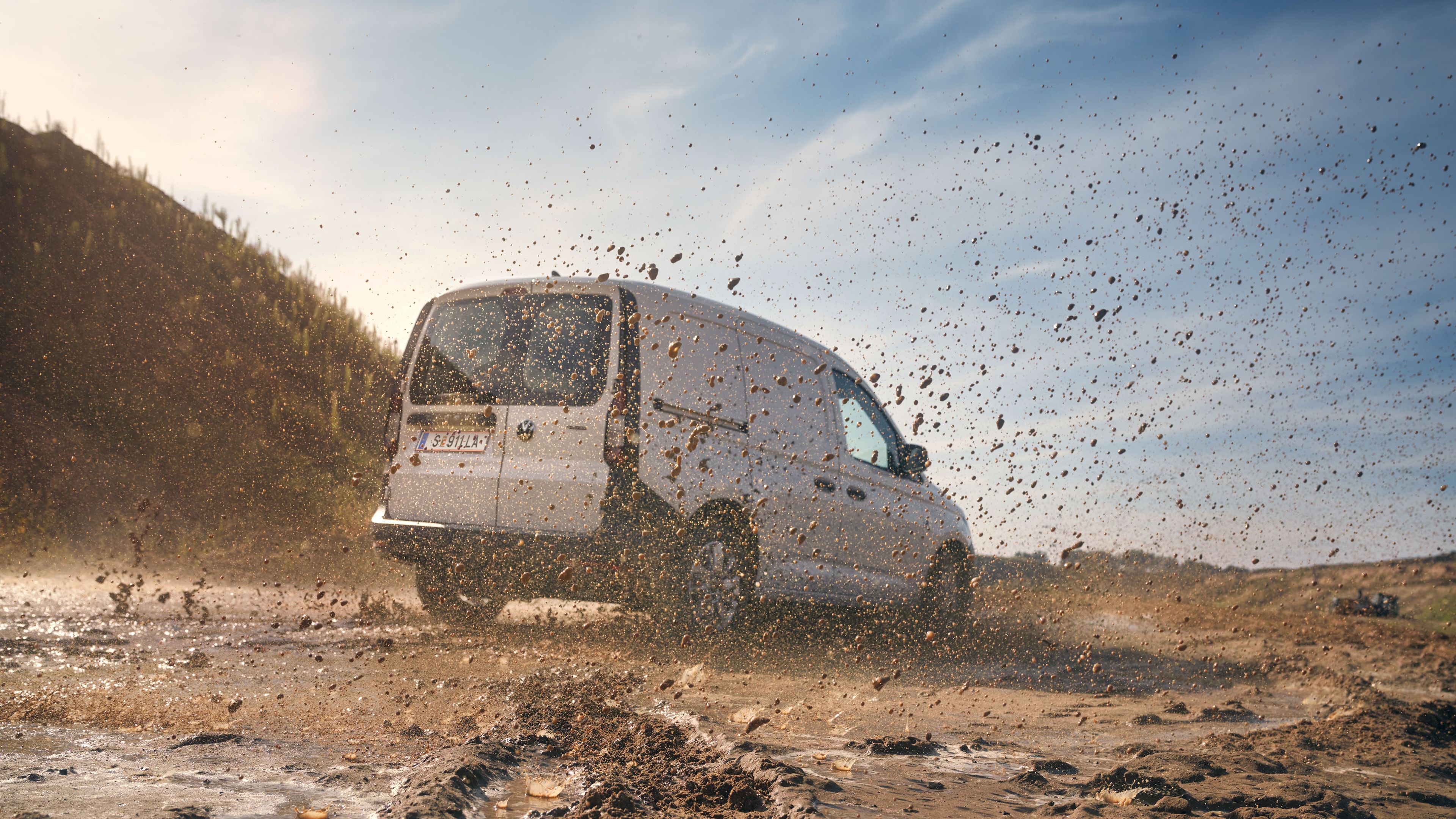 Ein weißer Caddy Kombi fährt dynamisch über schlammigen Untergrund. Das Wasser aus einer großen Pfütze spritzt hoch.